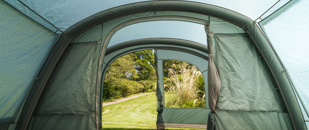 an image shot inside a tent showing the centre beam and open door with open window. scene seen through the tent is of a green grass field with paved path.