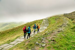 four friends walking up a path in the hills. The sky is grey and cloudy and the ground is wet and green. The people are wearing waterproof coats and trousers in an array of bright colours.