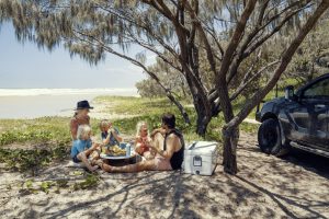 a family sat on sand under a shady branches tree eating lunch by the sea