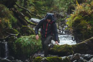 Man trekking in a rocky mountainous trail with a stream in the background. The man is climbing over a large rock underfoot in navy and red gear.